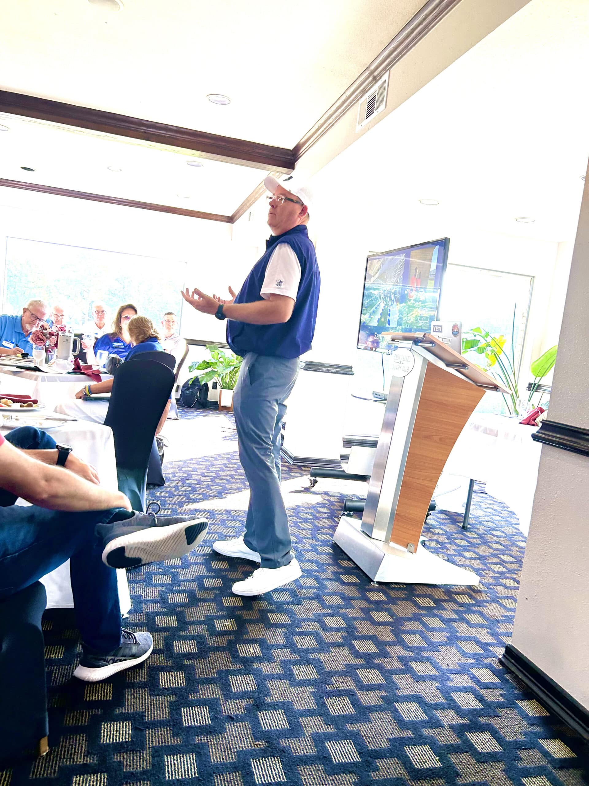 Men Serving God member delivering a presentation at a luncheon with attendees seated around tables and a screen showing visuals