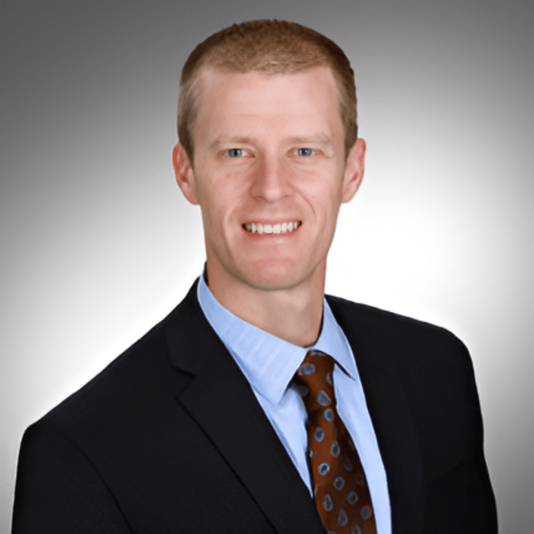 Professional headshot of a smiling man in a navy suit, light blue shirt, brown patterned tie, gray gradient background.