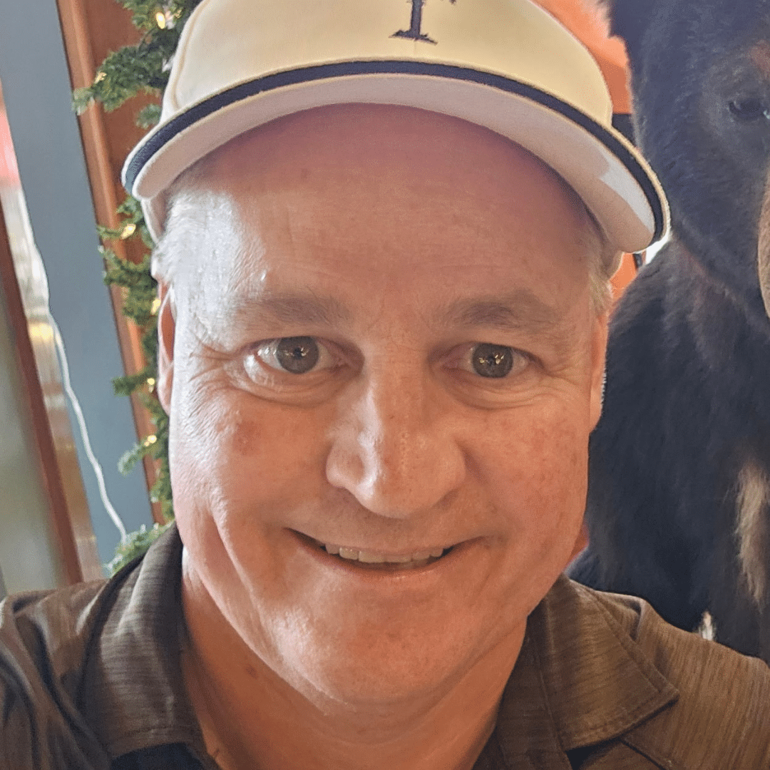 Smiling man wearing a light baseball cap taking a selfie, with a dark horse partially visible beside him.